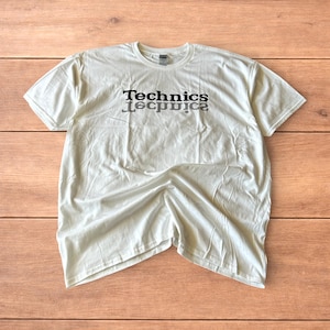 Könnte beinhalten: Ein hellfarbenes T-Shirt mit dem Wort "Technics" in Schwarz aufgedruckt, darunter ein Spiegelbild des Wortes. Das Shirt liegt flach auf einer Holzoberfläche.