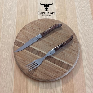 May include: A steak knife and fork set with wooden handles and stainless steel blades and tines, on a round wooden cutting board. The "Carnivore Kitchen" logo is above the board.