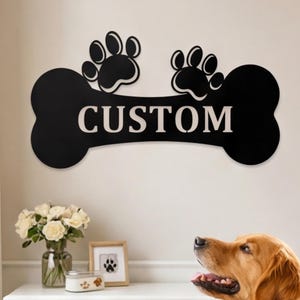 May include: A black metal dog bone-shaped sign with the word "CUSTOM" in white lettering. Two black paw prints are above the bone. A golden retriever looks up at the sign. The sign is a decorative item for pet owners.