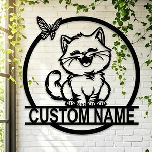 May include: Black metal cat wall art with a smiling cat inside a circle, a butterfly, and a horizontal bar with the text "CUSTOM NAME". The cat has a fluffy tail and is sitting. The background is a white brick wall.