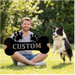 May include: A black bone-shaped sign with the word "CUSTOM" in a light color, and paw prints. A person is holding the sign while sitting on the grass with a black and white dog jumping nearby. The background is a green, leafy area.