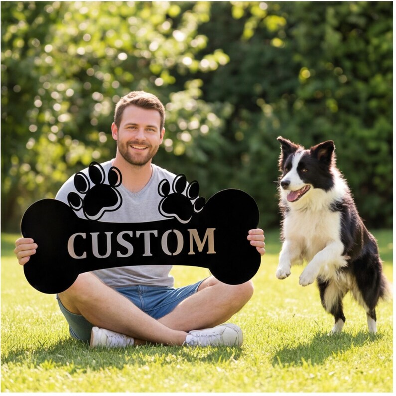 May include: A black bone-shaped sign with the word "CUSTOM" in a light color, and paw prints. A person is holding the sign while sitting on the grass with a black and white dog jumping nearby. The background is a green, leafy area.