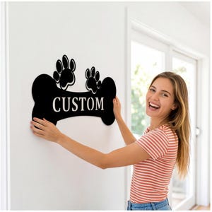 May include: Black bone-shaped sign with the word "CUSTOM" in white lettering. Two paw prints are above the word. The sign is being held up against a white wall.