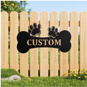 May include: Black metal dog bone-shaped sign with the word "CUSTOM" cut out. Two paw prints are above the bone. The sign is mounted on a wooden fence with green grass and yellow flowers in the foreground.