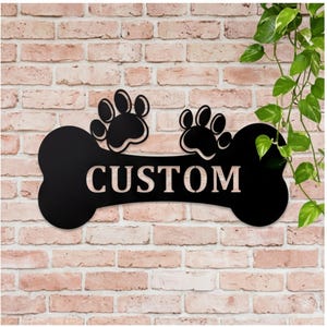 May include: Black metal dog bone-shaped sign with two paw prints and the word "CUSTOM" in white. The sign is mounted on a brick wall, with green leafy vines in the background.