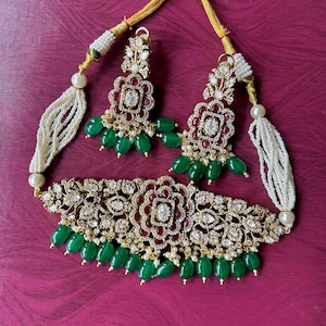 May include: A jewelry set consisting of a choker necklace and matching earrings. The pieces feature gold-tone metal, clear crystals, red stones, and dangling green beads. The necklace includes multi-strand white seed bead accents on each side.