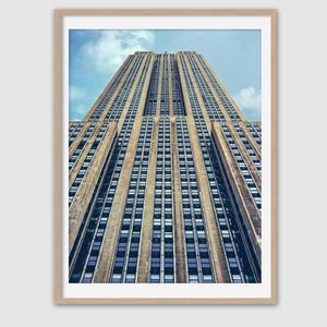 May include: An upward view of the Empire State Building in New York City, framed in a light wood-colored frame. The building's facade is a light brown color with many windows. The sky is a light blue with some clouds.