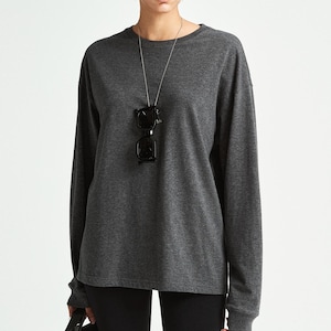 May include: A dark charcoal gray long-sleeve crewneck shirt paired with black fitted knee-length shorts. A thin silver chain necklace holds a pair of black rectangular sunglasses. A black tote bag is held at the side of the shorts.