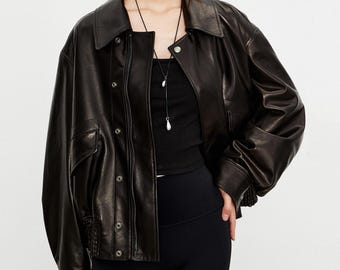 May include: A black leather jacket with a wide collar, front zipper, and snap button closure. The jacket features long sleeves with ribbed cuffs, two front pockets, and a smooth finish. It is worn over a black top and black leggings.