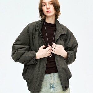 May include: A dark olive green bomber jacket with a high collar and a zipper closure. The jacket is paired with a dark brown shirt and light blue jeans. A silver necklace is visible.