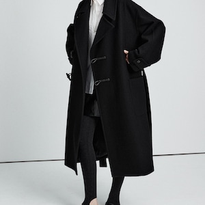 May include: A long, black wool coat with a classic design. The coat features a notched collar, two front pockets, and a double-breasted closure. The model is wearing a white shirt, black leggings, and black shoes. The text "WOMEN'S COLLECTION" is visible.