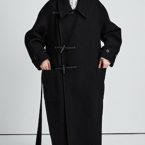 May include: A black wool-blend duffle coat featuring a wide collar, long sleeves with buttoned cuffs, and two large front patch pockets. The coat is secured with two toggle closures and includes a matching fabric belt hanging from the side.