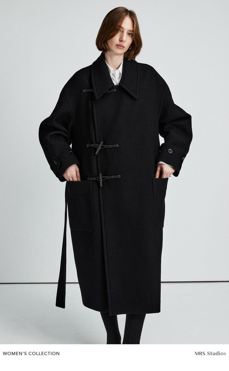 May include: A black wool-blend duffle coat featuring a wide collar, long sleeves with buttoned cuffs, and two large front patch pockets. The coat is secured with two toggle closures and includes a matching fabric belt hanging from the side.