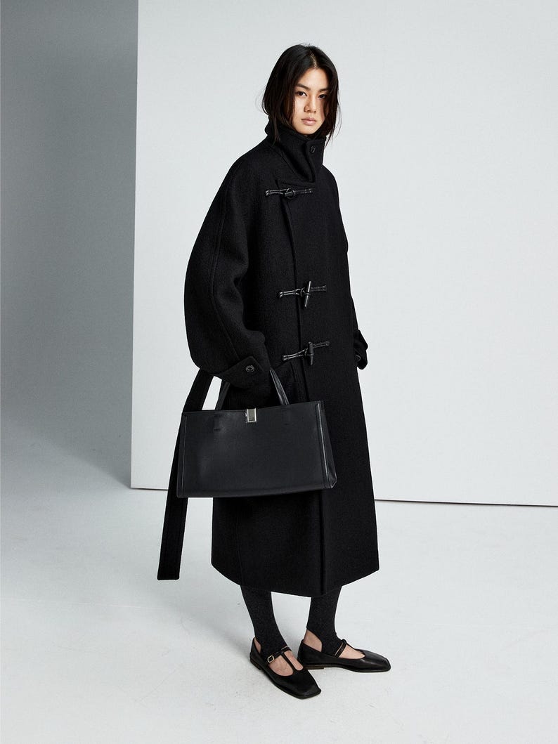 May include: A black wool-blend duffle coat featuring a high collar, toggle closures, and long sleeves. The coat is paired with a black rectangular leather handbag with a silver-tone metal clasp, black stirrup leggings, and black flat Mary Jane shoes.