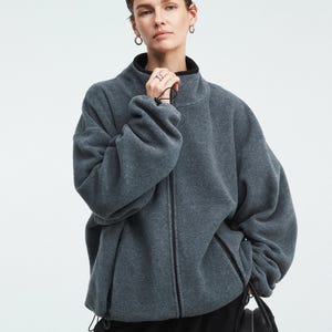May include: A gray fleece jacket with a high collar and a full-length zipper. The jacket has a black trim on the collar and cuffs. The model is wearing black pants and holding a black bag. The jacket appears to be made of a soft, warm material.