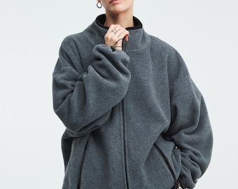 Puede incluir: Una chaqueta de forro polar gris carbón con cuello alto, cremallera frontal negra completa y dos bolsillos laterales con cremalleras negras. La chaqueta tiene mangas largas con puños elásticos y un dobladillo ajustable con cordón.