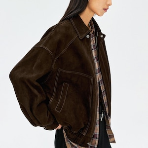 Könnte beinhalten: Dunkelbraune Wildlederjacke mit Kragen und zwei Vordertaschen. Die Jacke hat eine lockere Passform mit geripptem Saum und Bündchen. Die Person trägt ein kariertes Hemd darunter.