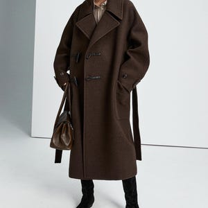 May include: A dark brown wool-blend coat featuring a wide collar, toggle closures, and a matching waist belt. The coat is paired with black suede boots and a dark brown leather handbag. The text "WOMEN'S COLLECTION" and "MRS Studios" is visible.