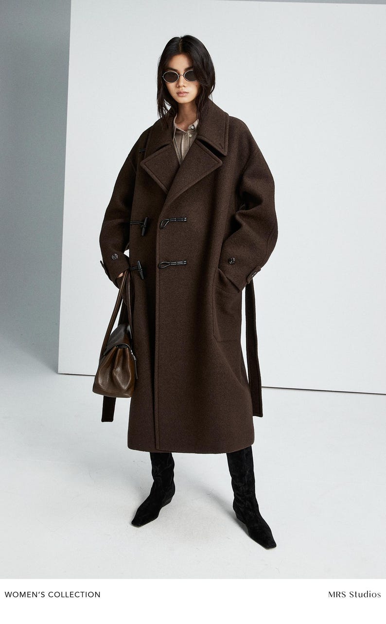 May include: A dark brown wool-blend coat featuring a wide collar, toggle closures, and a matching waist belt. The coat is paired with black suede boots and a dark brown leather handbag. The text "WOMEN'S COLLECTION" and "MRS Studios" is visible.