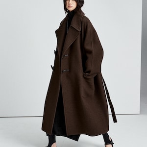 May include: A long, dark brown wool coat featuring a wide collar, dropped shoulders, and toggle closures. The coat is worn over a black turtleneck sweater, a black skirt, and black flat shoes with a single strap across the top of the foot.