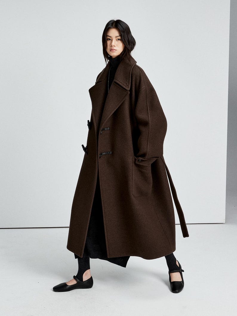 May include: A long, dark brown wool coat featuring a wide collar, dropped shoulders, and toggle closures. The coat is worn over a black turtleneck sweater, a black skirt, and black flat shoes with a single strap across the top of the foot.
