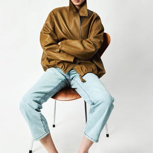 May include: An oversized tan textured jacket with a high collar and a front zipper closure, paired with light blue denim jeans and black shoes. The jacket features long sleeves and a loose fit, while the jeans have a straight-leg cut and cropped hem.