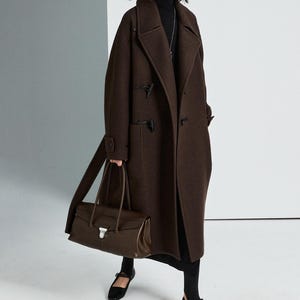 May include: A dark brown wool duffle coat with toggle closures and large patch pockets, worn over a black turtleneck and skirt. A dark brown leather handbag with a silver metal clasp is carried alongside black flat shoes with a strap.