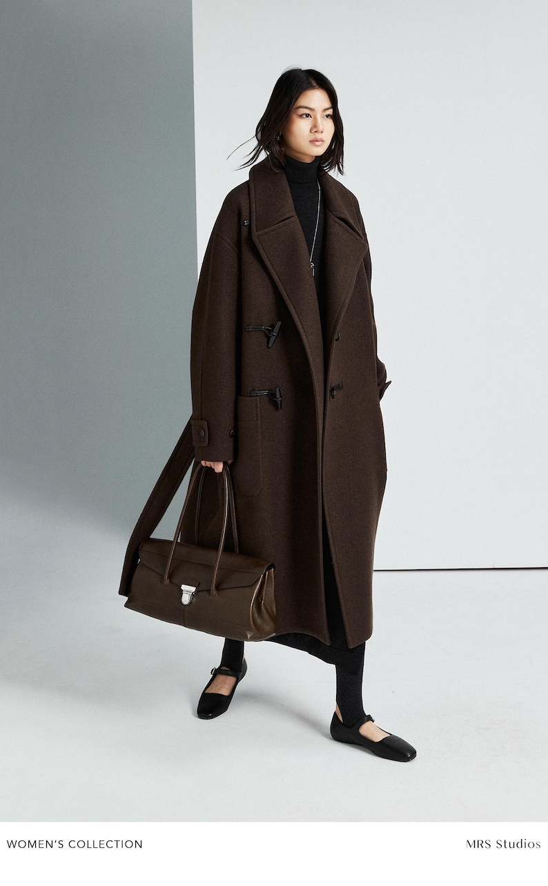 May include: A dark brown wool duffle coat with toggle closures and large patch pockets, worn over a black turtleneck and skirt. A dark brown leather handbag with a silver metal clasp is carried alongside black flat shoes with a strap.