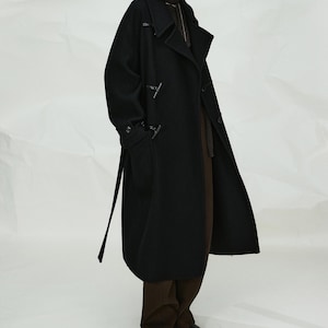 May include: A long black wool-blend duffle coat featuring a wide collar, toggle closures, and long sleeves. The coat is worn over dark brown trousers and black shoes. The text "WOMEN'S COLLECTION" and "MRS Studios" appears at the bottom of the image.