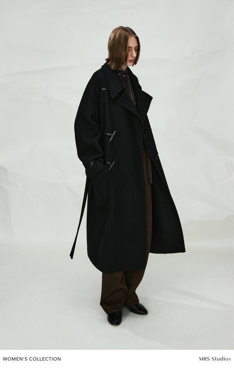 May include: A long black wool-blend duffle coat featuring a wide collar, toggle closures, and long sleeves. The coat is worn over dark brown trousers and black shoes. The text "WOMEN'S COLLECTION" and "MRS Studios" appears at the bottom of the image.