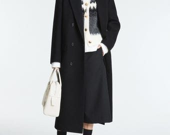 Può includere: Una donna indossa un cappotto nero doppiopetto, una camicia bianca, un gilet lavorato a maglia fantasia e una gonna nera. Tiene in mano una borsa bianca e indossa calzini e scarpe neri. Collezione donna.