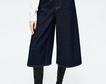 May include: Dark wash denim culottes with a black belt and black boots. The culottes have a wide leg design and are paired with a white long-sleeve top. The outfit is set against a white background.