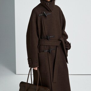 May include: A dark brown wool coat featuring a high collar, long sleeves with buttoned cuffs, and a front closure with three black toggle fasteners. The coat includes a matching tie belt at the waist and two large patch pockets on the front.
