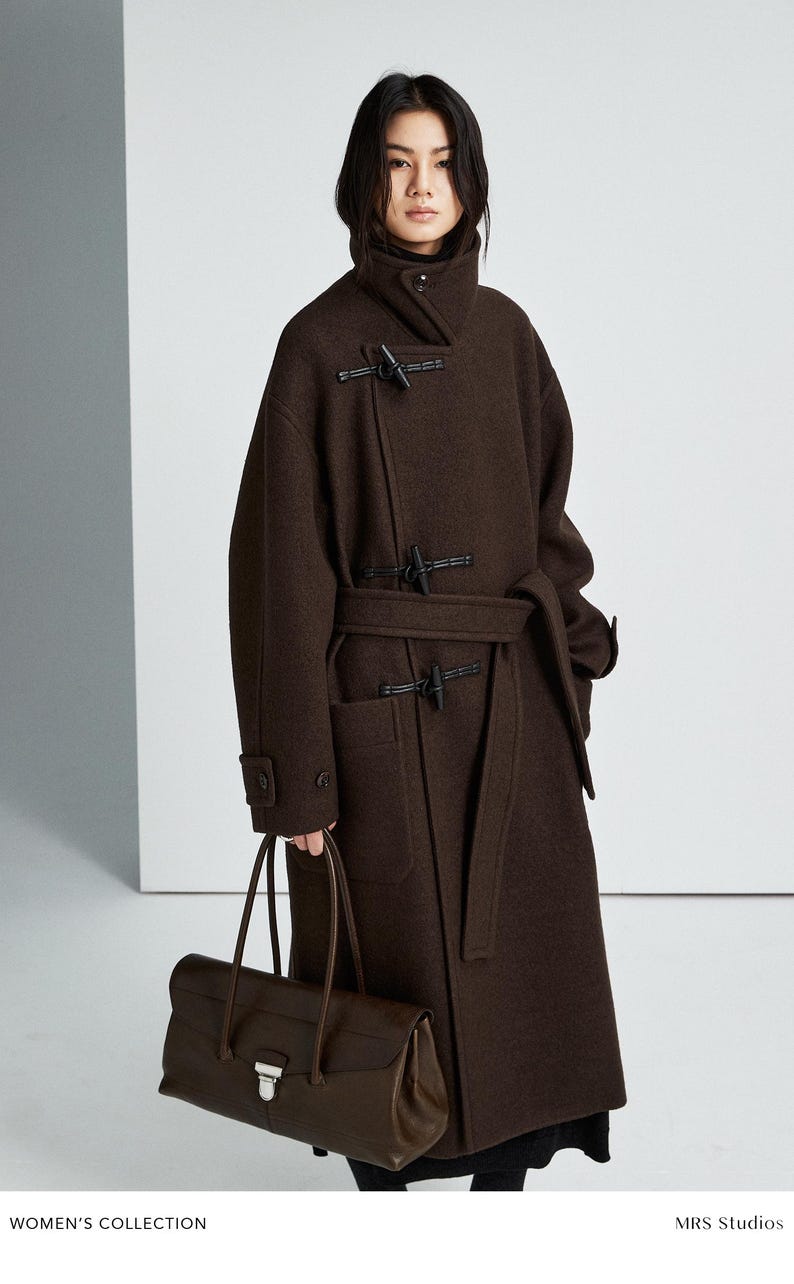 May include: A dark brown wool coat featuring a high collar, long sleeves with buttoned cuffs, and a front closure with three black toggle fasteners. The coat includes a matching tie belt at the waist and two large patch pockets on the front.
