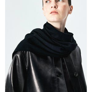May include: A black knit scarf wrapped around the neck, worn over a black leather jacket with a button-down front. The jacket features a smooth, reflective surface and a structured collar, while the scarf has a soft, textured appearance.