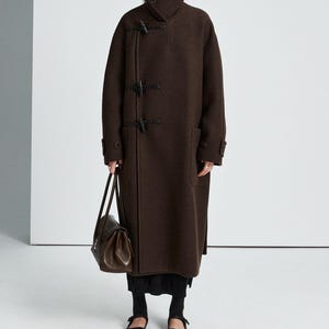 May include: A dark brown wool-blend coat featuring a high collar, long sleeves, and three black toggle closures on the front. The coat is paired with a dark brown leather handbag, a black skirt, black socks, and black Mary Jane style shoes.