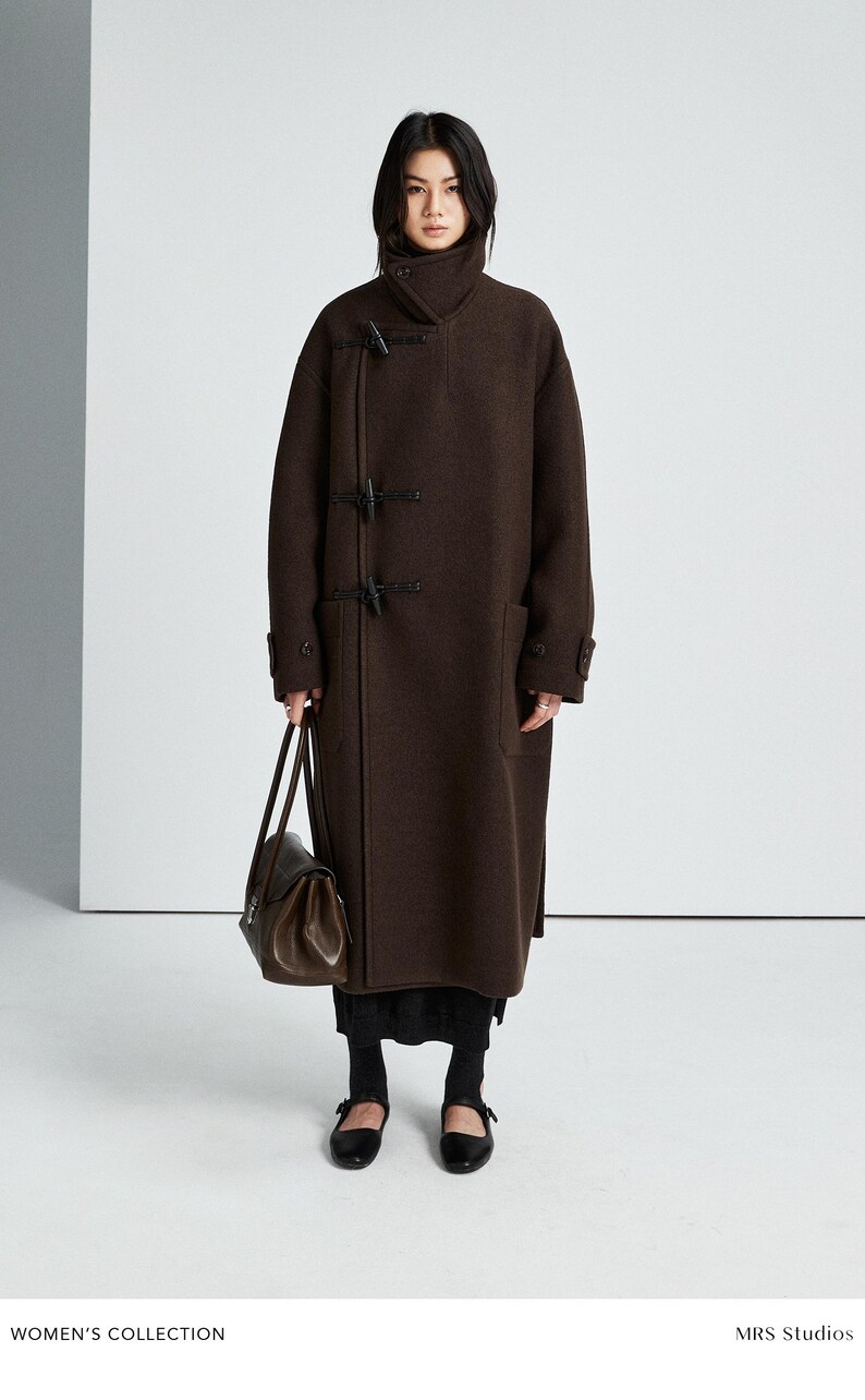 May include: A dark brown wool-blend coat featuring a high collar, long sleeves, and three black toggle closures on the front. The coat is paired with a dark brown leather handbag, a black skirt, black socks, and black Mary Jane style shoes.