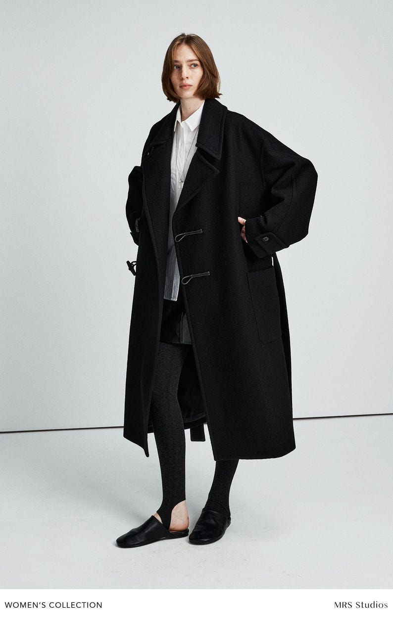 May include: A long, black wool coat with a classic design. The coat features a notched collar, two front pockets, and a double-breasted closure. The model is wearing a white shirt, black leggings, and black shoes. The text "WOMEN'S COLLECTION" is visible.