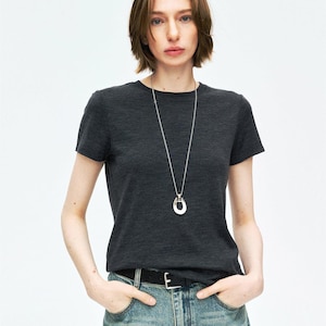 May include: A dark charcoal gray short-sleeved t-shirt paired with a long silver-toned chain necklace featuring a horseshoe-shaped pendant. The outfit includes light blue denim jeans secured with a black belt that has a silver-toned buckle.