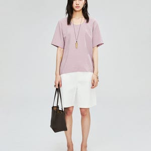 May include: A woman wearing a mauve short-sleeved top and white shorts. She is carrying a dark brown tote bag and wearing a long necklace with a gold pendant. She is also wearing sandals.