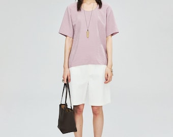 May include: A woman wearing a mauve short-sleeved top and white shorts. She is carrying a dark brown tote bag and wearing a long necklace with a gold pendant. She is also wearing sandals.