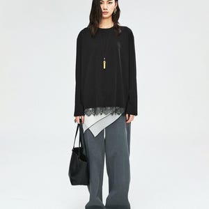 May include: A woman wearing a black long-sleeve top with a white and black lace trim, paired with gray wide-leg pants. She carries a black tote bag and wears black sandals. A gold pendant necklace is visible.