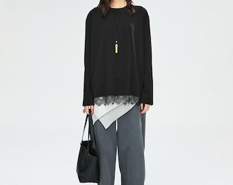 May include: A woman wearing a black long-sleeve top with a white and black lace trim, paired with gray wide-leg pants. She carries a black tote bag and wears black sandals. A gold pendant necklace is visible.