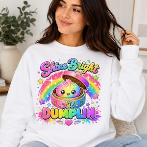 May include: A white sweatshirt featuring a colorful graphic of a smiling rainbow dumpling inside a bamboo steamer. The text reads Shine Bright Lil DUMPLIN, surrounded by a vibrant rainbow, stars, and paint splatters in pink, blue, and yellow hues.