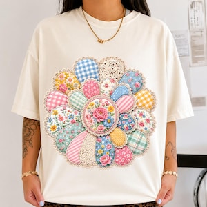 Puede incluir: Una camiseta de manga corta color crema con un gran gráfico circular en la parte delantera. El diseño es una flor compuesta por varios parches de tela estampada, incluyendo cuadros vichy, estampados florales y texturas de encaje.