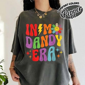 Op de afbeelding: Donkergrijs t-shirt met de kleurrijke retro-opdruk "IN MY DANDY ERA". Het ontwerp bevat een gele bliksemschicht, een lachende bloem en sterren. Het t-shirt heeft korte mouwen en een relaxte pasvorm.