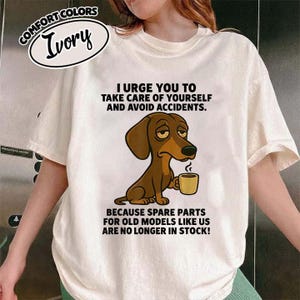 Puede incluir: Camiseta de color marfil con el texto "I urge you to take care of yourself and avoid accidents." Se muestra un perro salchicha de dibujos animados sosteniendo una taza de café. El texto de abajo dice "Because spare parts for old models like us are no longer in stock!"