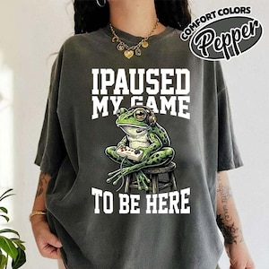 Puede incluir: Camiseta gris oscuro con un diseño de una rana verde con auriculares y un mando de videojuegos. El texto blanco dice "I PAUSED MY GAME TO BE HERE". La camiseta tiene mangas cortas y un corte holgado. También se ve el texto "COMFORT COLORS Pepper".