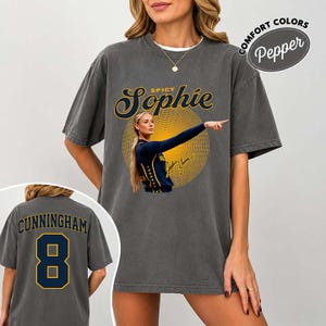 May include: A charcoal gray short-sleeve t-shirt featuring a graphic of a basketball player in a blue uniform. The text reads Spicy Sophie above the player and Cunningham 8 on the back. A circular yellow sunburst pattern sits behind the player.