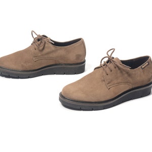 Puede incluir: Un par de zapatos marrones de ante con cordones y suelas negras. Los zapatos tienen una plataforma ligeramente elevada y el nombre de la marca "Mobils" es visible en el zapato derecho.